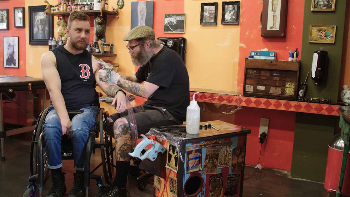 Jack receiving a tattoo on his left arm from a guy at a tattoo parlour. 