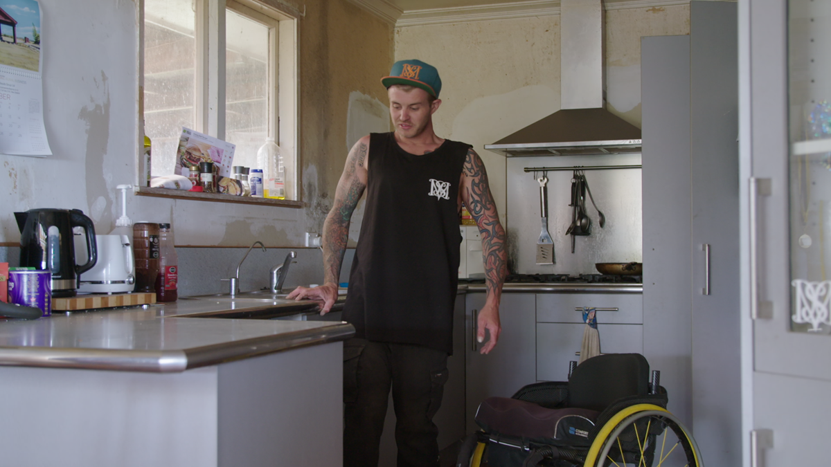 Scott stands next to his wheelchair holding his balance on the kitchen bench.
