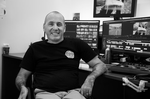 A man in a wheelchair wears a black t-shirt and sits in front of a computer with three displays.
